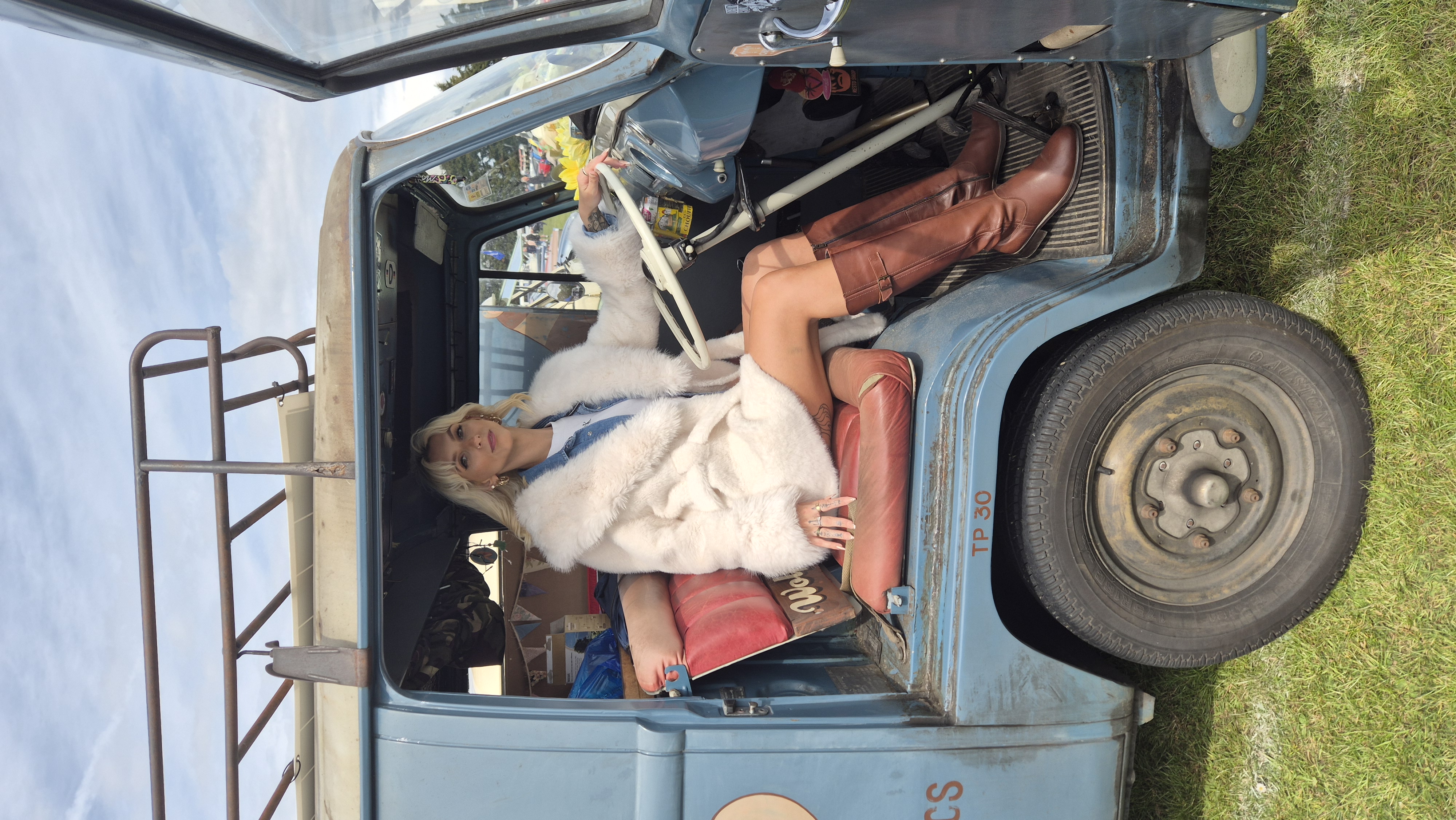 Emma in a cream faux-fur coat and tan riding boots in a vintage blue camper van
